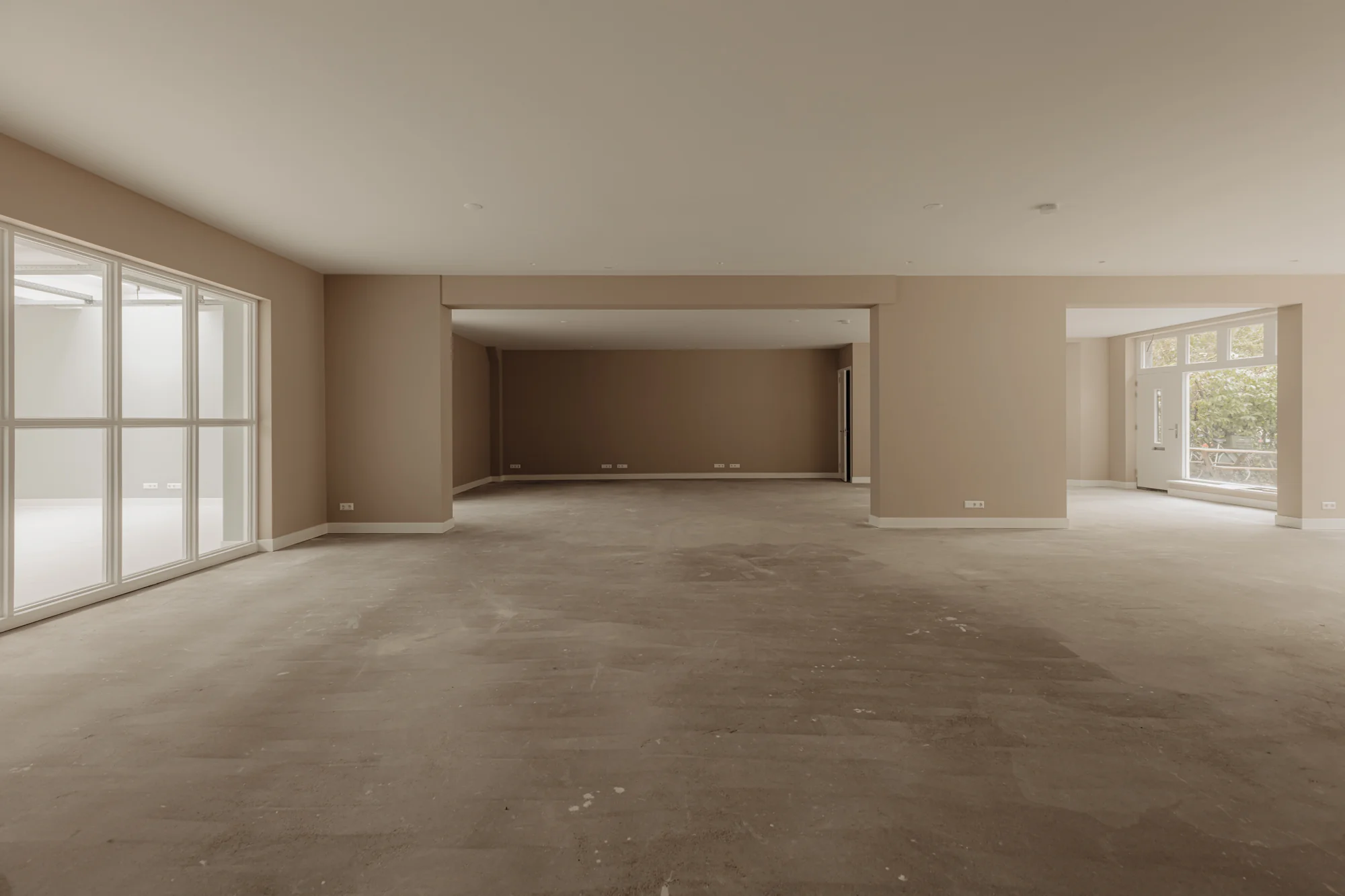 Ruime, leegstaande kamer met beige muren, grote ramen en betonnen vloer aan de Henri Polaklaan.